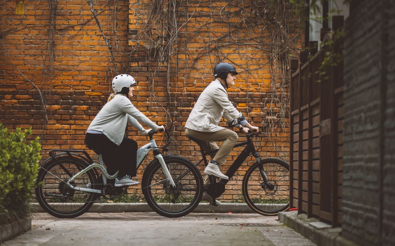 Couple cycling on E-bikes through a city lane with a classic brick wall backdrop.
